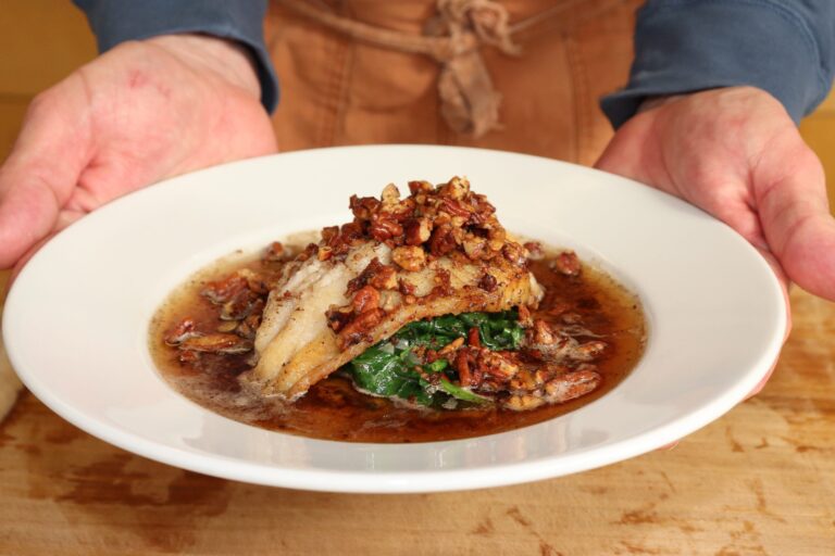 Person presenting a plated pecan-crusted catfish fillet over sautéed spinach with a brown butter pecan sauce.