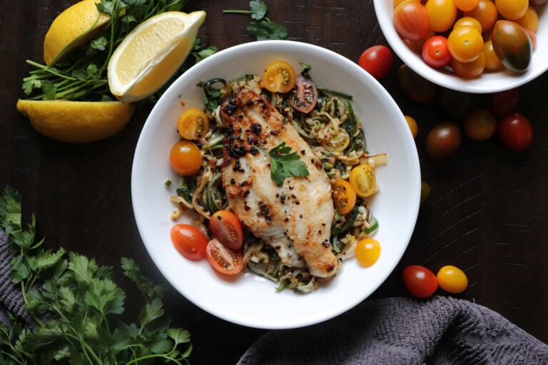 Seared catfish fillet plated over sautéed vegetable noodles with halved cherry tomatoes, garnished with parsley and lemon wedges.