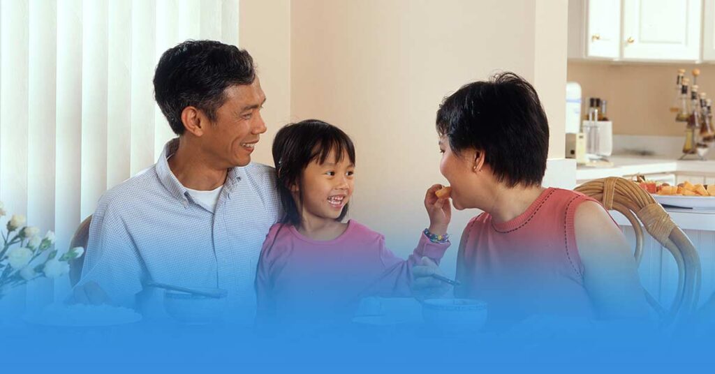 A father and daughter smiling at the table while a mother tastes a bite of food, in a home dining setting.
