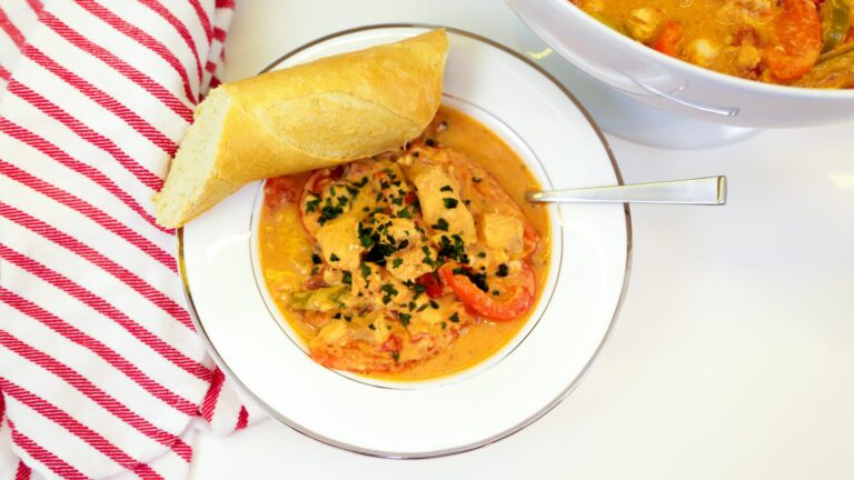 Bowl of catfish and vegetable stew garnished with parsley, served with a slice of French bread and a spoon, alongside a serving dish of the same stew.