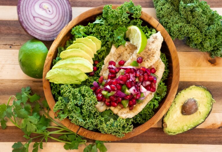 Overhead view of a wooden bowl filled with a seared catfish fillet topped with pomegranate salsa, served over kale with avocado and lime, surrounded by fresh ingredients.
