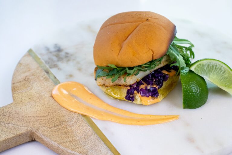 Grilled catfish sandwich on a toasted bun with arugula, purple cabbage slaw, and a swoosh of spicy sauce, served with lime wedges.