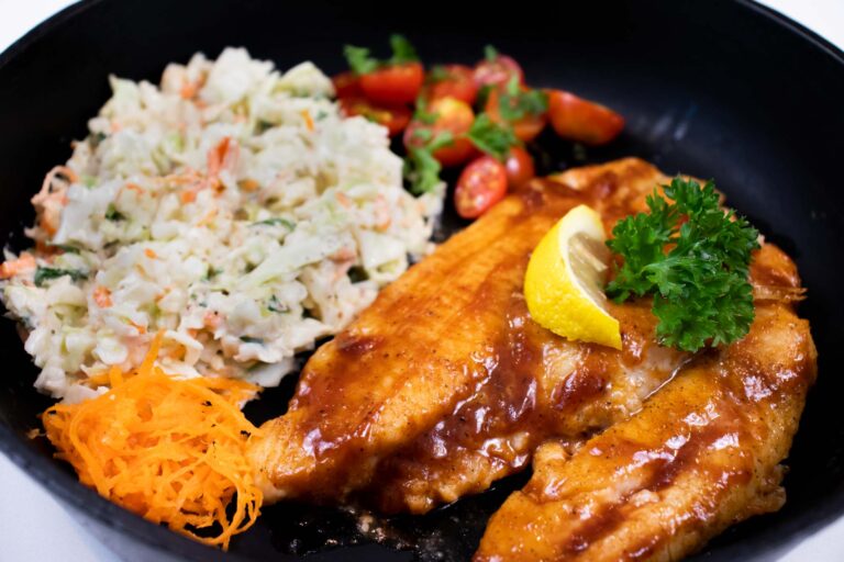 Barbecue-glazed catfish fillets served with coleslaw, shredded carrots, and cherry tomatoes, garnished with lemon and parsley.