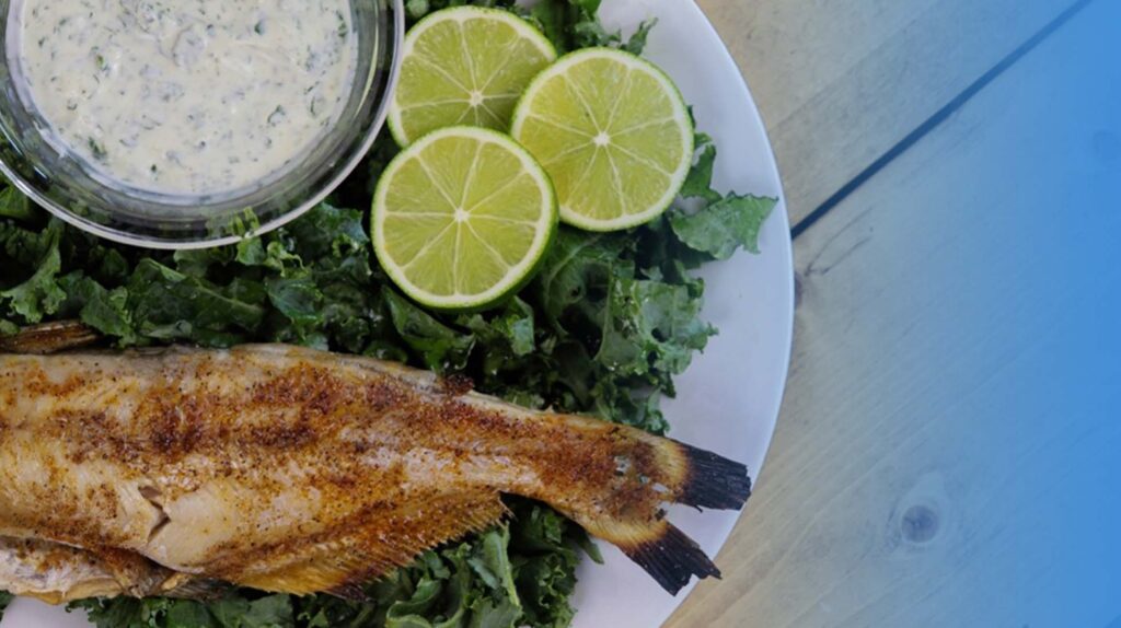 Overhead view of a seasoned whole catfish on a bed of kale, served with lime slices and a creamy herb dip.