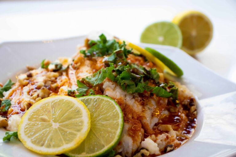 Close-up of a catfish fillet topped with chopped nuts and a savory sauce, garnished with kale, lemon, and lime slices.