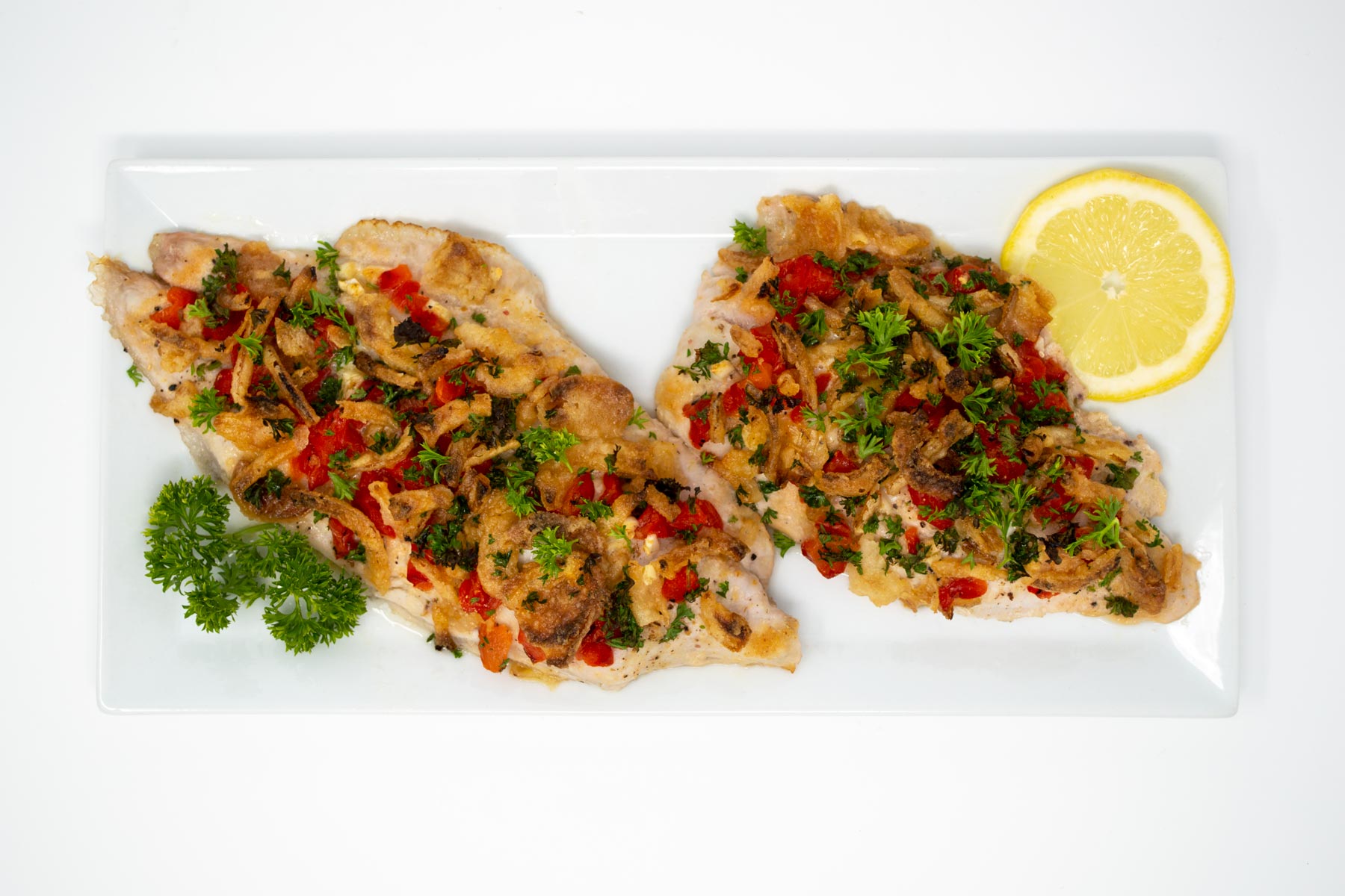 Two grilled catfish fillets topped with crispy onions, diced red pepper, and parsley, garnished with a lemon slice.