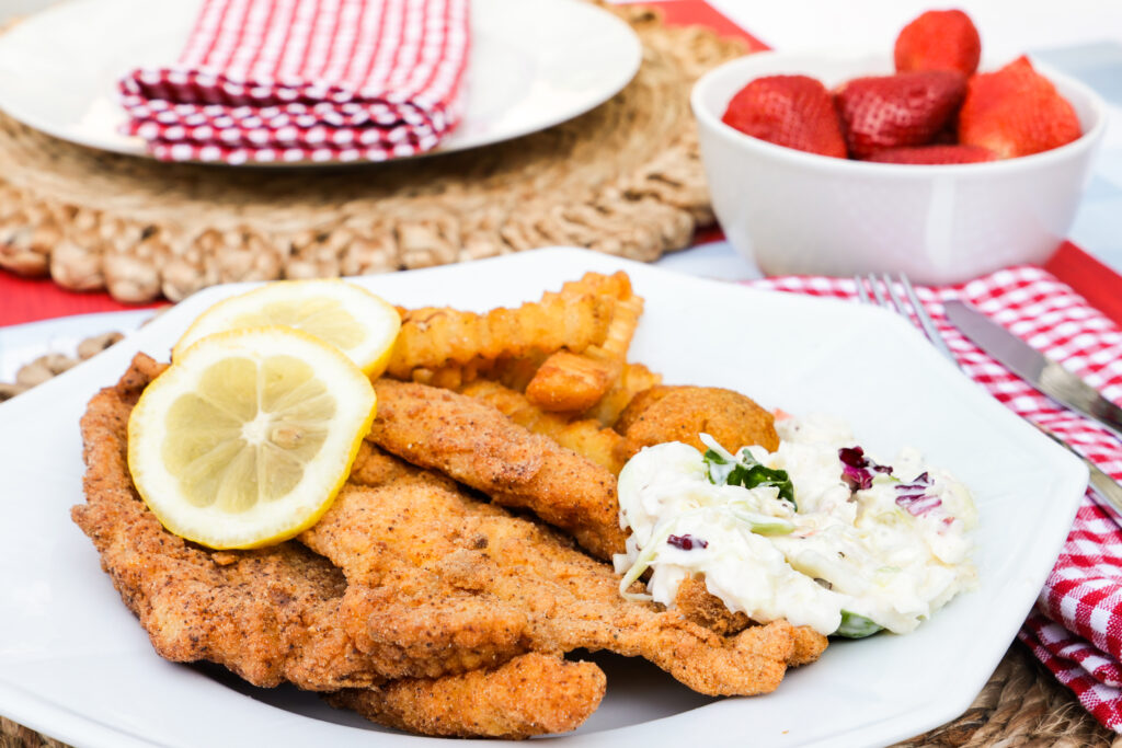 Fried catfish fillets served with crinkle-cut fries, coleslaw, and lemon slices at an outdoor table set with a red gingham napkin and a bowl of strawberries.