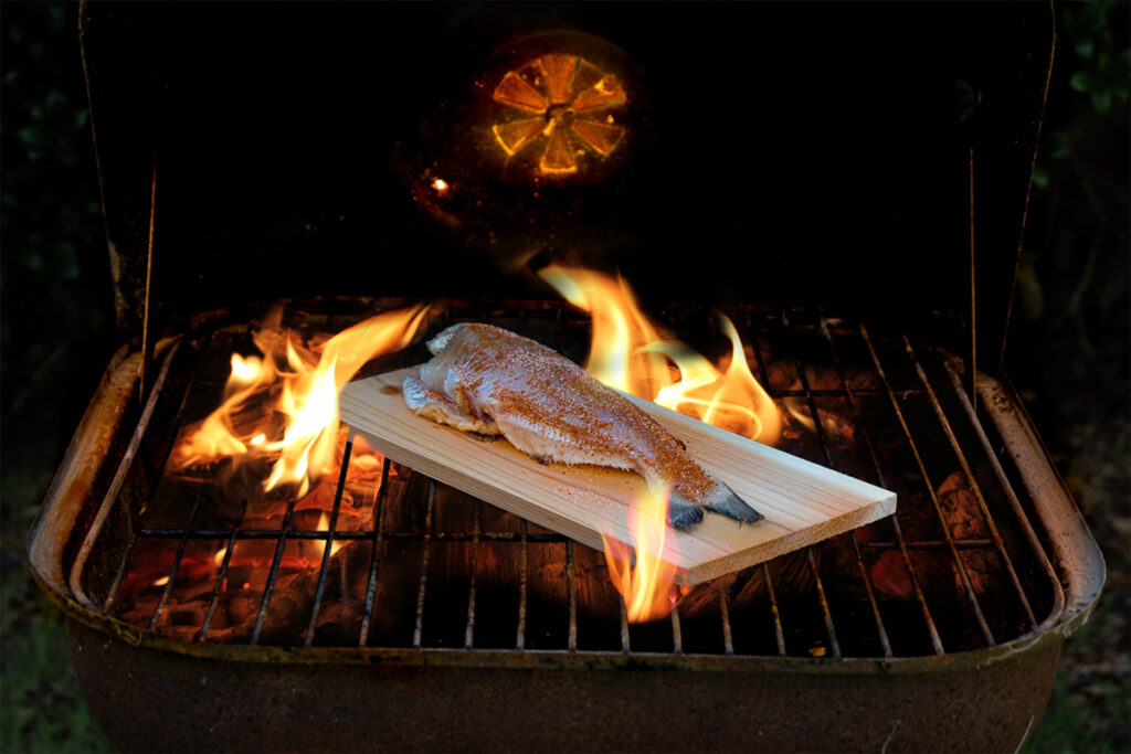 Whole seasoned catfish grilling on a cedar plank over open flames on an outdoor grill.