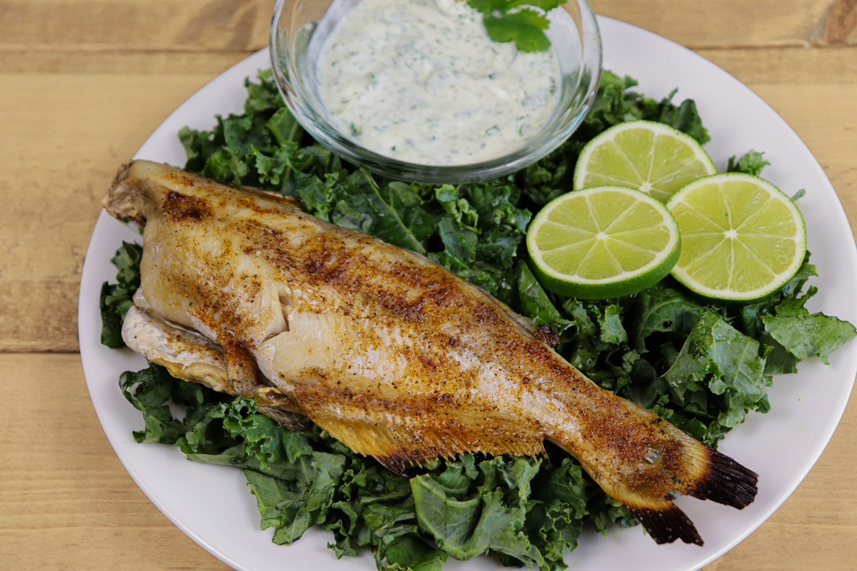 Whole seasoned catfish served on a bed of kale with lime slices and a creamy herb dipping sauce.